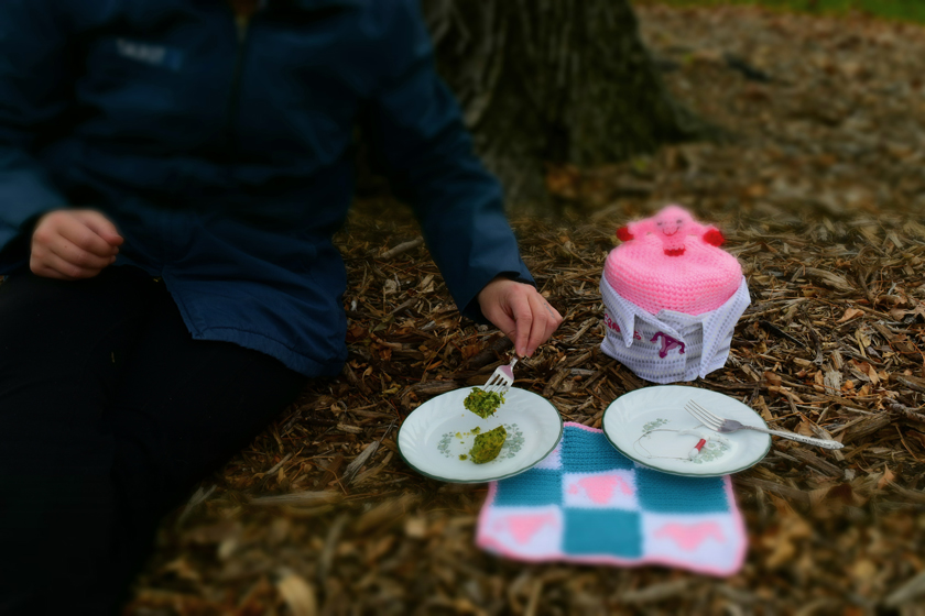 DSC_0174 Chris having a picnic in the park with their uterus in a Tupperware. The uterus has a ceramic IUD on their plate and is wearing a jacket and a beanie hat where the beanie is a uterus.