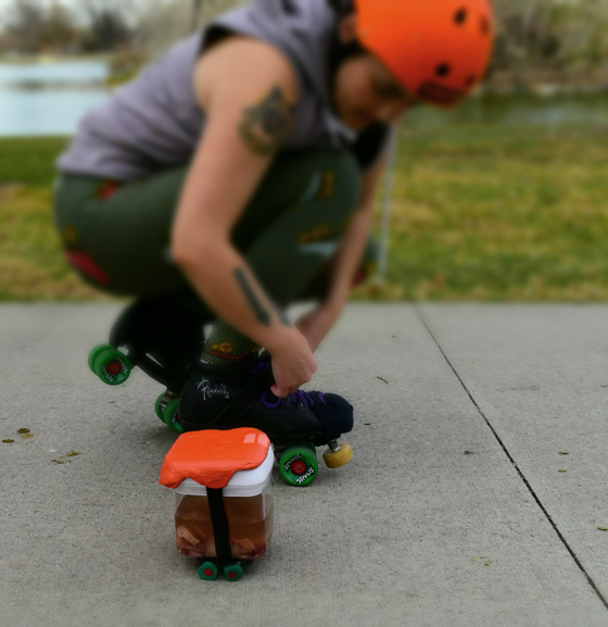 DSC_0183 A uterus in a Tupperware is wearing an orange ceramic helmet and skates. Behind them Chris is putting on their skates and is wearing a matching orange helmet.
